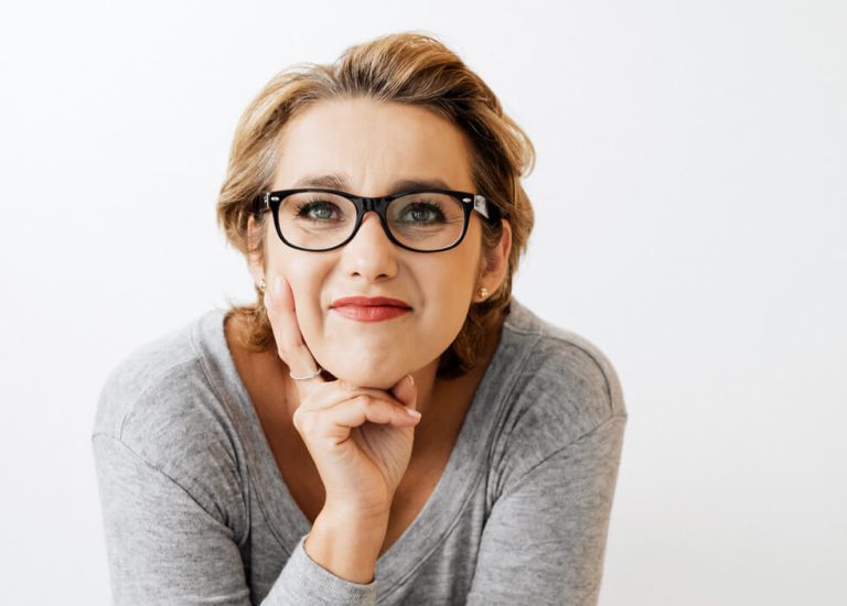 A woman with short light brown hair and black glasses rests her chin on her hand, wearing a light gray top, against a plain white background.