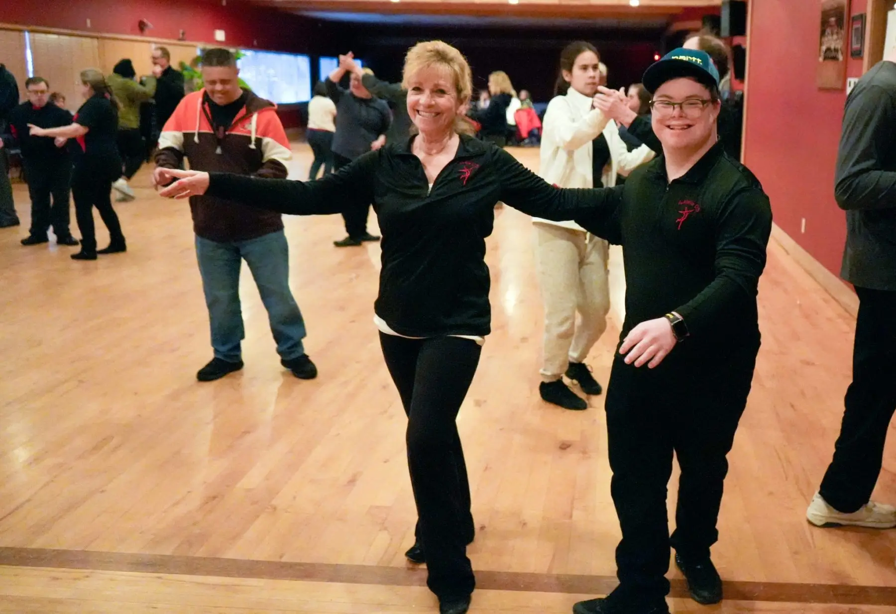 A woman and a young man dance together in a studio with wooden floors, while other people dance in the background.