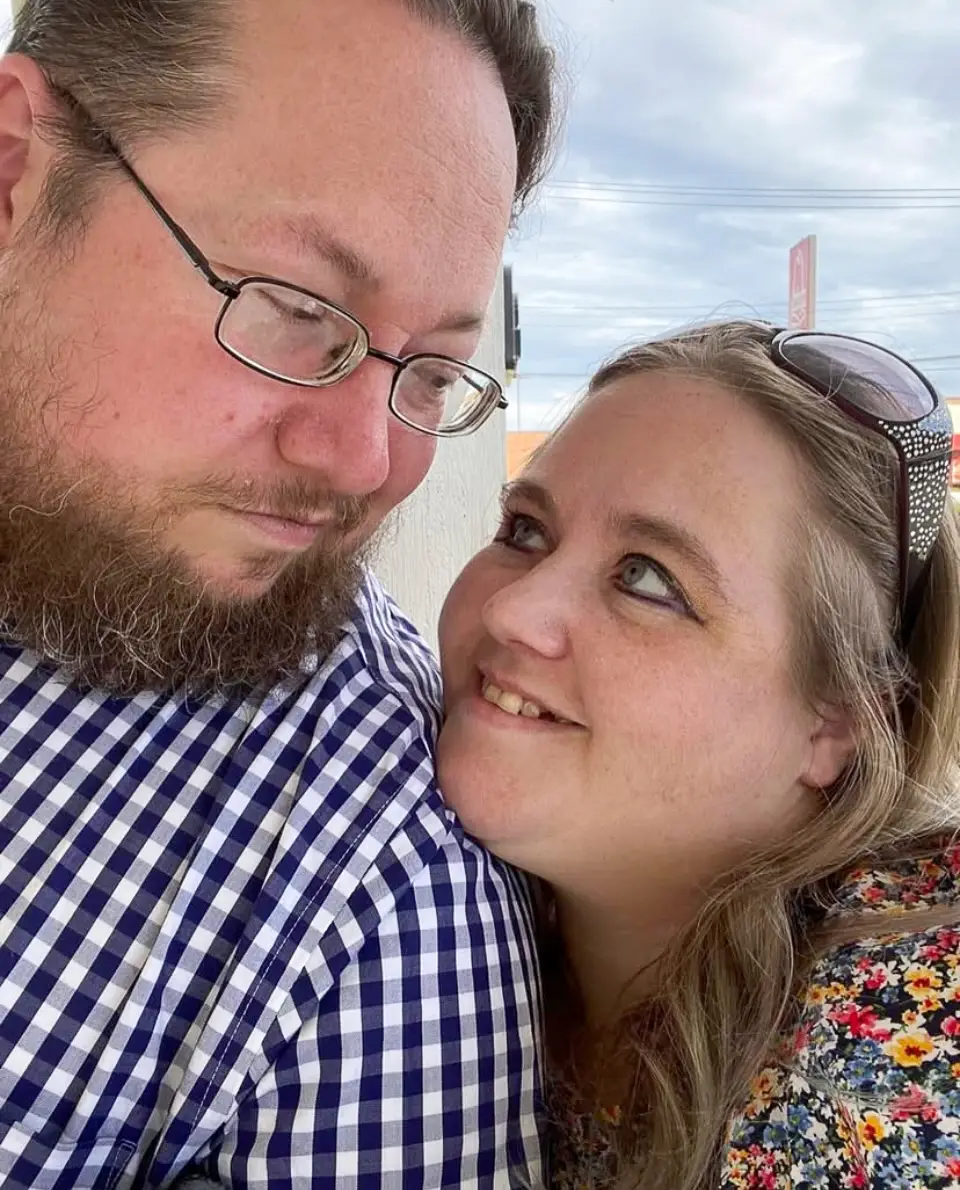 A man in a checkered shirt looks down at a woman smiling up at him. She wears a floral top and sunglasses on her head. They are outdoors with a cloudy sky in the background.
