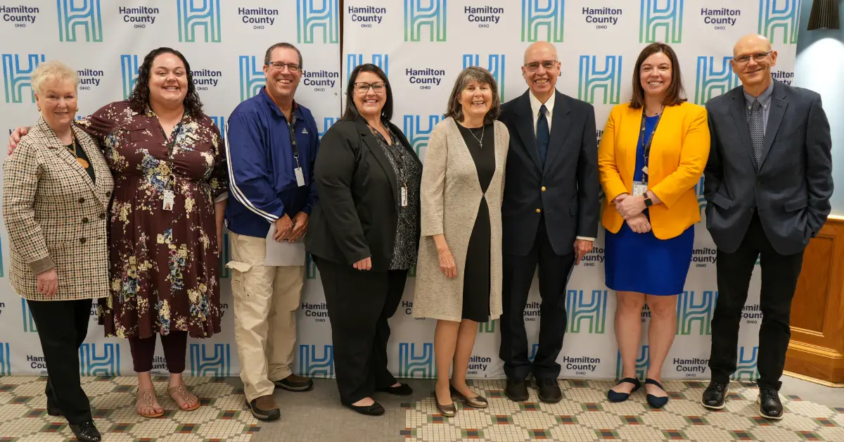 Eight adults stand in a row, smiling at the camera, in front of a backdrop with the Hamilton County logo, celebrating "Jack Boehnlein Day."